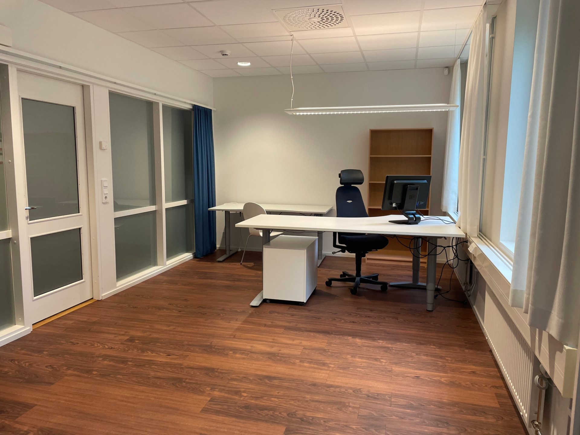 Större kontorsrum med två fönster, skrivbord, mörkblå kontorsstol, bokhylla och glasparti med dörr
