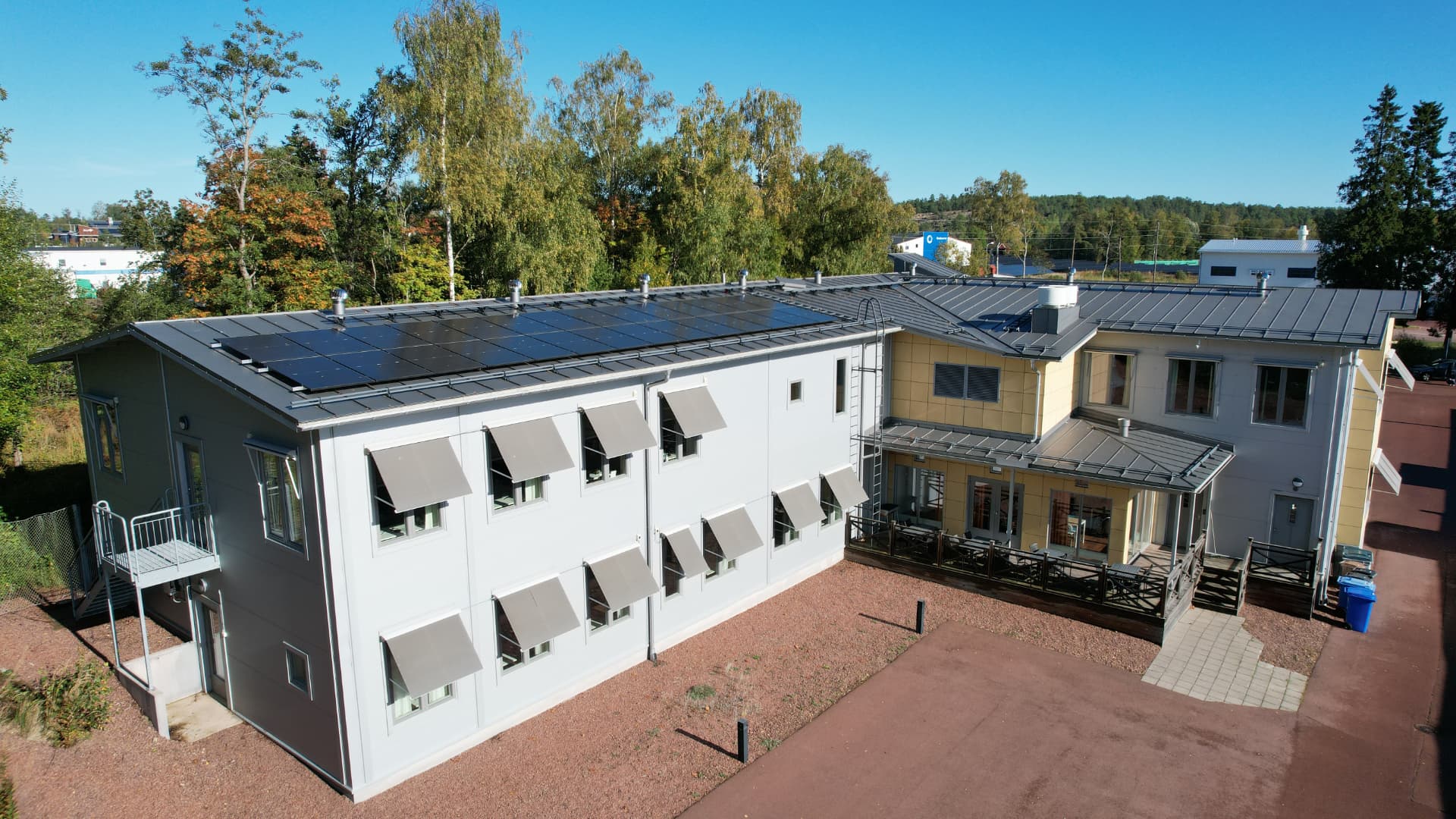 Flygfoto av fastigheten på Godbyvägen 2 i Mariehamn med solpaneler på taket, terrass vid entrén och de stora parkeringsytorna framför byggnaden
