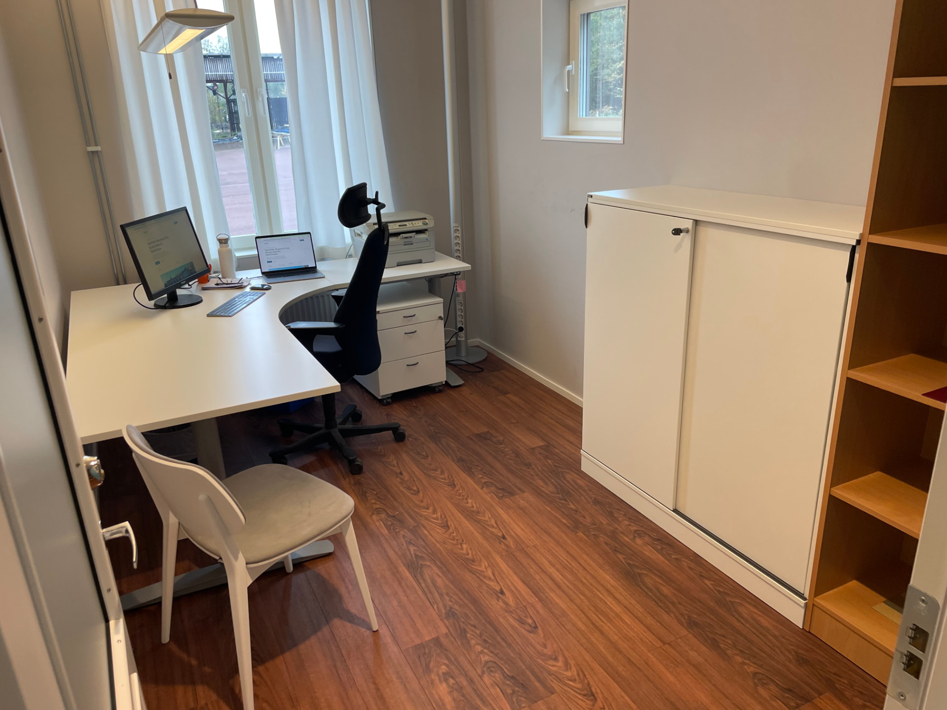 Enskilt kontorsrum med vinkelskrivbord, kontorsstol, skåp och fönster med gardiner